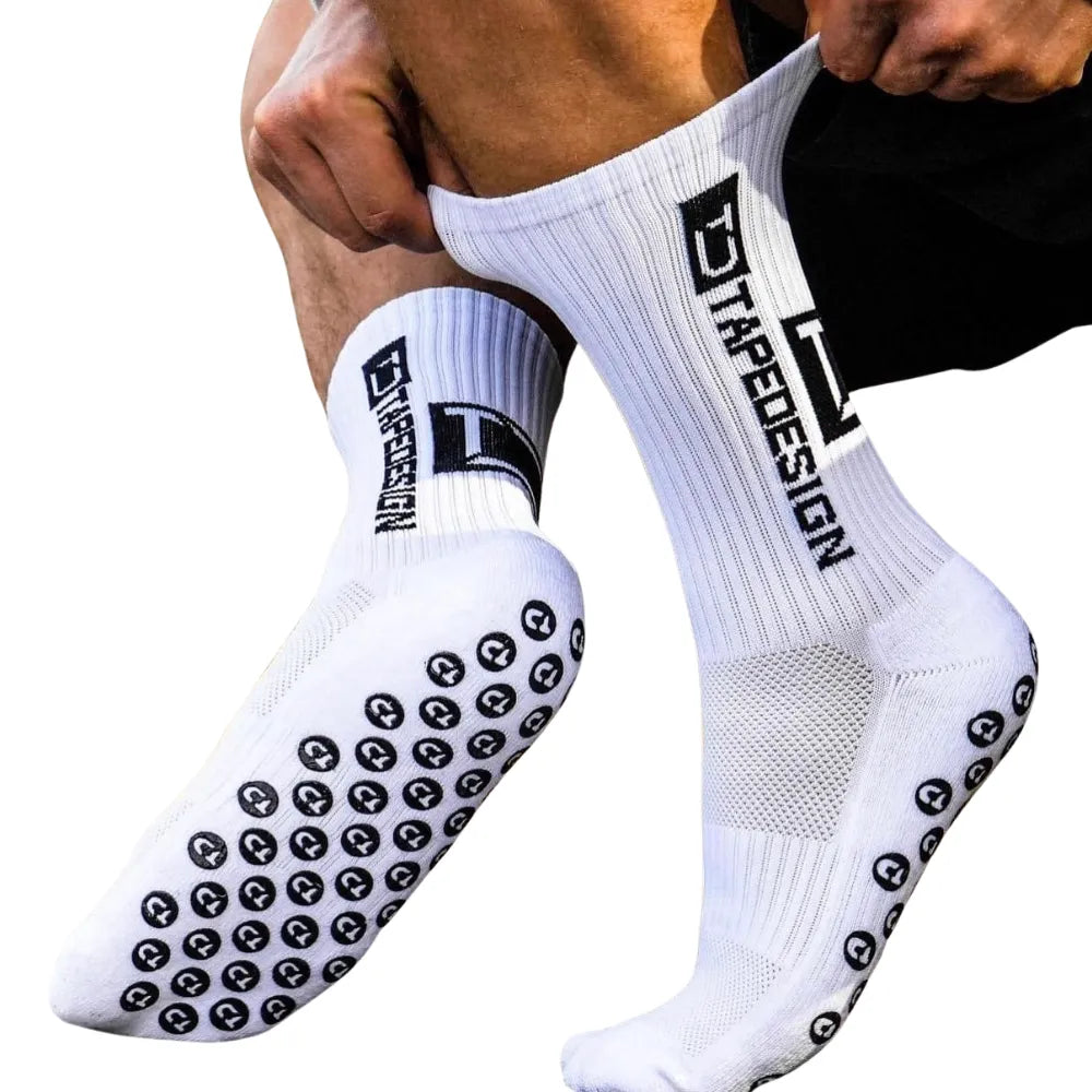 Man putting on Tapedesign non-slip socks, one foot raised on tiptoe while holding the top of the sock, the other foot showing the anti-slip sole.